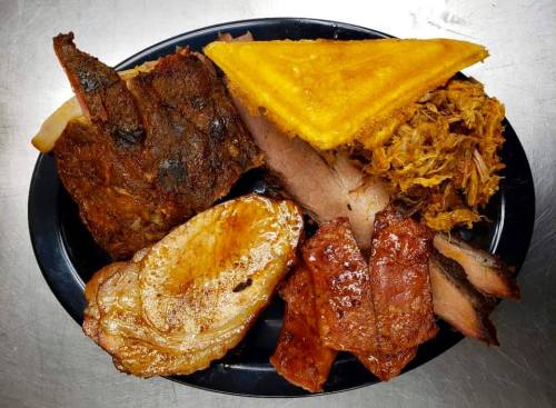 A platter of assorted meats including ribs, pulled pork, brisket, and cornbread, served on a black plate.