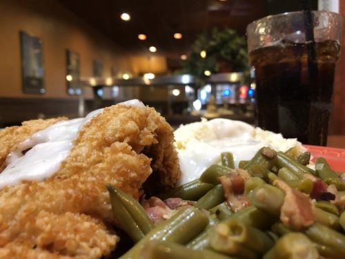 A plate of fried chicken with gravy, mashed potatoes, and green beans, alongside a glass of cola in a restaurant setting.