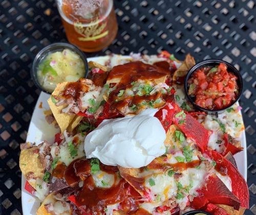 A plate of loaded nachos topped with cheese, sour cream, barbecue sauce, and sides of salsa and guacamole, with a beer.