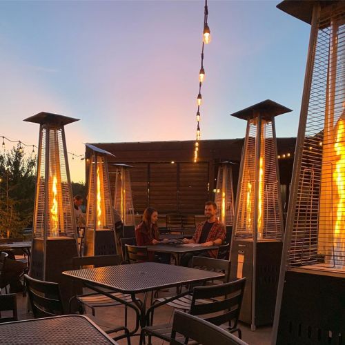 A cozy outdoor dining area with two people at a table, surrounded by patio heaters and string lights at sunset.