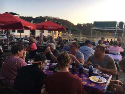 A lively outdoor gathering by the water, with people enjoying food and drinks under red umbrellas at sunset.