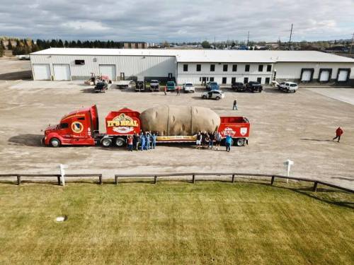 A large potato on a truck, surrounded by people, with a warehouse in the background on a cloudy day.