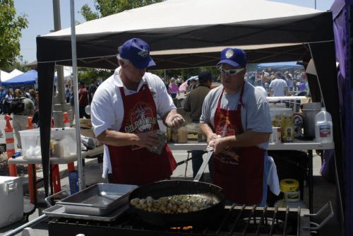 Two men in aprons cooking at a food stall, surrounded by a busy outdoor event with tents and people in the background.