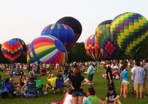 A crowd gathers on a grassy field to watch colorful hot air balloons being inflated for a festival.
