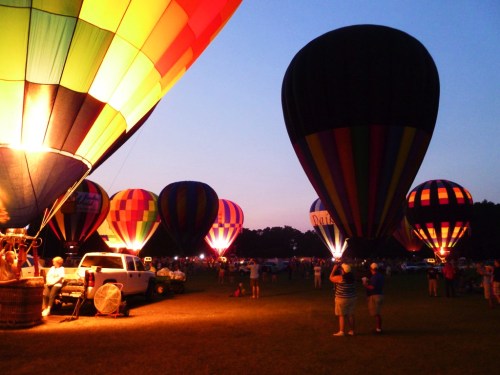 Colorful hot air balloons illuminated at dusk, with spectators gathered on a grassy field.