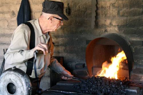 An elderly man works at a forge, tending to glowing coals and flames in a rustic blacksmith shop.