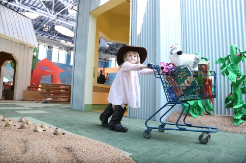 A child in a hat pushes a shopping cart filled with toys in a colorful indoor play area.