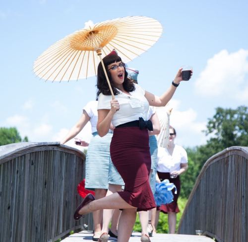 A woman in a vintage outfit holds a parasol and a drink, joyfully posing on a wooden bridge with friends in the background.