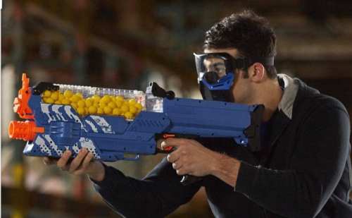 A person wearing a mask aims a blue toy blaster filled with yellow balls in an indoor setting.