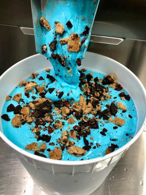 A large bucket filled with bright blue ice cream topped with cookie crumbles and chocolate pieces.