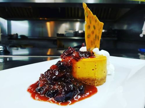 A slice of cheesecake topped with berry compote, whipped cream, and a crispy caramel shard on a white plate.