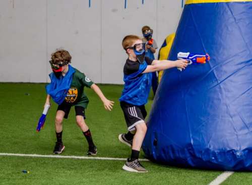 Two children in blue vests and masks play with toy blasters in an indoor arena, aiming at a large inflatable barrier.
