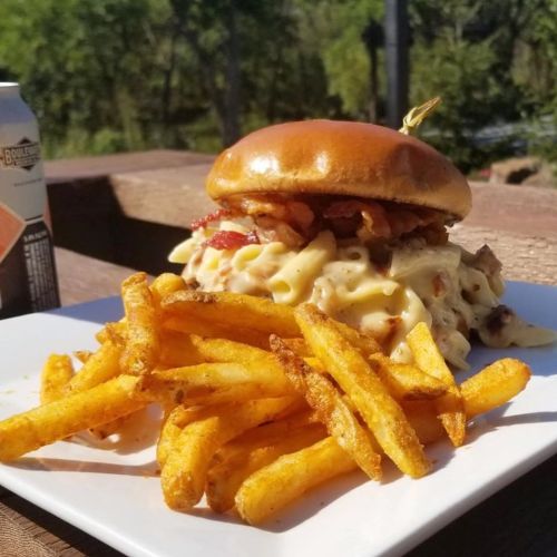 A hearty burger topped with bacon and creamy pasta, served with golden fries on a white plate outdoors.