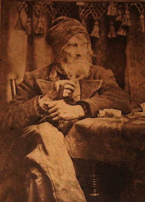 A sepia-toned portrait of an elderly man with a long beard, wearing a hat, seated at a table with a contemplative expression.