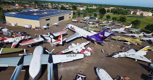 Aerial view of various aircraft displayed outside a building, with people exploring the exhibits in a sunny setting.
