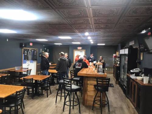A cozy café interior with wooden tables, bar seating, and a group of people chatting near the counter.