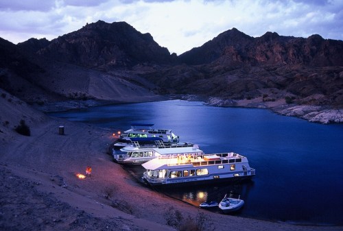 A serene lakeside scene at dusk with houseboats moored along a sandy shore, surrounded by mountains.