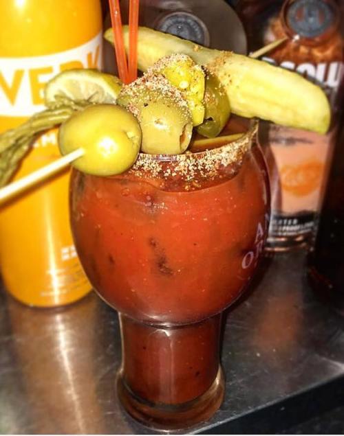 A vibrant Bloody Mary garnished with olives, pickles, and a lime wedge, set against a backdrop of liquor bottles.