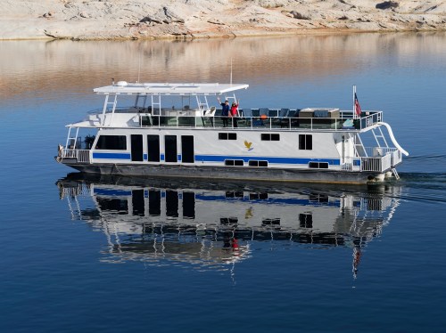A houseboat glides across calm water, reflecting its image against the serene landscape.