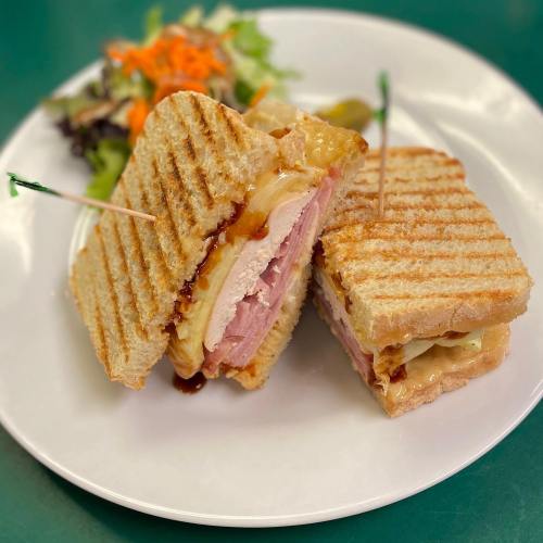 A grilled sandwich with layers of chicken, ham, and cheese, served with a side salad on a white plate.