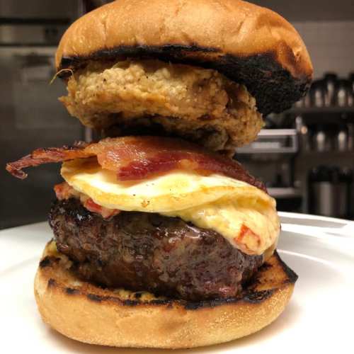 A towering burger with a beef patty, fried egg, bacon, and a crispy chicken fillet, all in a toasted bun.