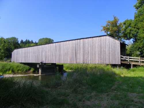 A wooden covered bridge spans a small river, surrounded by lush greenery and a clear blue sky.