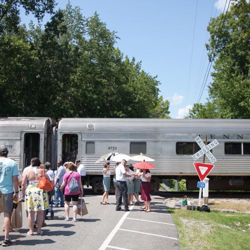 A crowd gathers near a train at a crossing, with trees and blue sky in the background. Some people hold umbrellas.