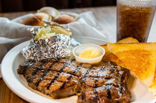 A plate with a grilled steak, baked potato in foil, butter, toast, and a glass of soda in the background.