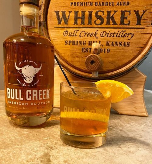 A bottle of Bull Creek American Bourbon next to a glass of whiskey garnished with an orange slice.