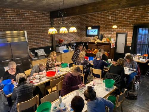 A group of people engaged in a crafting activity around tables in a cozy, well-lit room with brick walls.