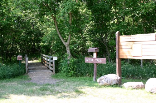 A nature trail entrance with wooden signs and a small bridge surrounded by lush greenery.