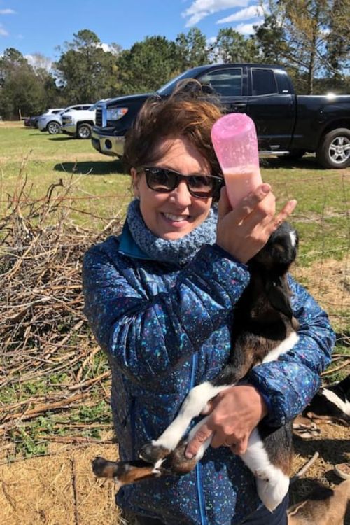 A woman in sunglasses holds a baby goat while feeding it from a pink bottle, smiling in a sunny outdoor setting.