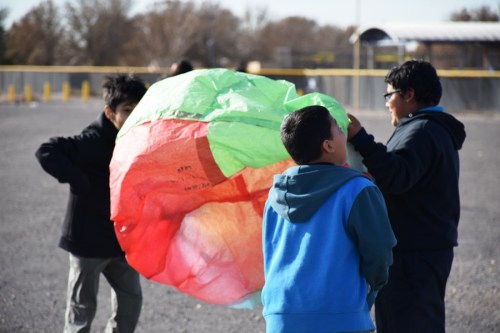 Children gather around a large, colorful balloon, preparing to launch it outdoors on a sunny day.