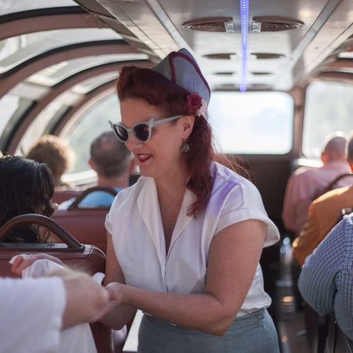 A woman in vintage attire serves passengers on a train, wearing sunglasses and a stylish hat, with a cheerful expression.