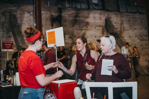 A vendor hands a sample to two women at a bustling event, with a rustic backdrop and various booths in view.