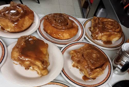 A tray of six large, gooey cinnamon rolls on plates, topped with a rich caramel glaze.