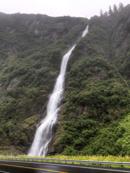 A cascading waterfall flows down a lush, green mountainside, with a road visible in the foreground.