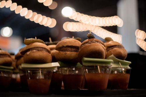 A close-up of mini burgers topped with pickles, arranged on a table with blurred lights in the background.