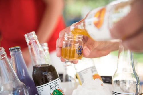 A hand pours a golden drink from a bottle into a small glass, surrounded by various colorful beverage bottles.