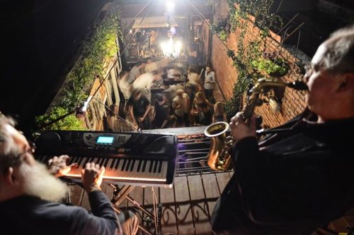 A musician plays keyboard while another plays saxophone at a lively rooftop party with people dancing below.