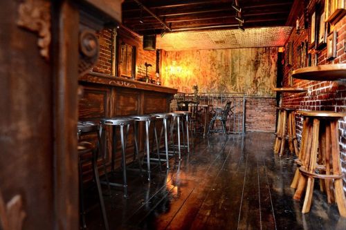 A rustic bar interior with wooden floors, brick walls, and high stools, creating a warm, inviting atmosphere.
