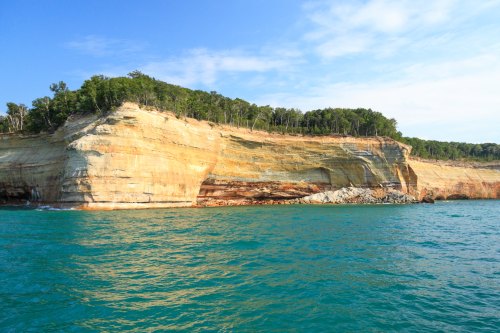 A scenic view of a rocky cliff with lush greenery above, rising from clear blue water under a bright sky.