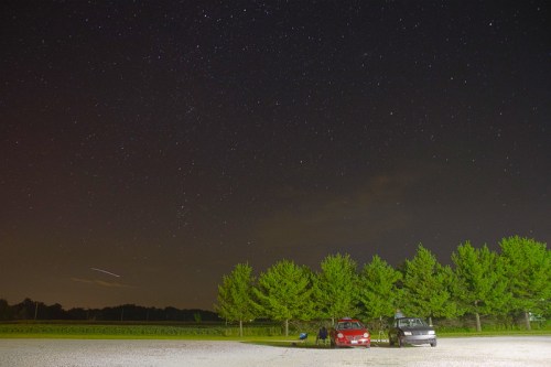 Two cars parked under a starry night sky, with trees in the background and a hint of clouds.