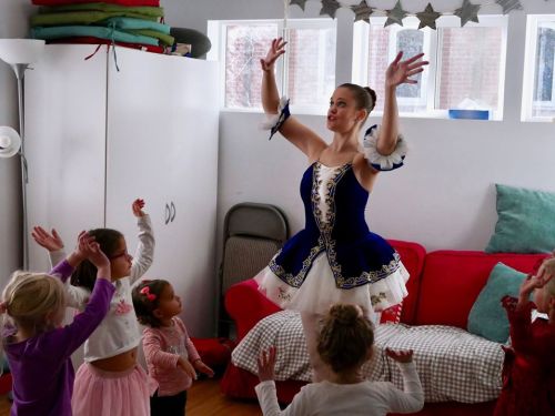 A ballet instructor in a blue costume demonstrates a pose to a group of young children in a bright, cozy room.