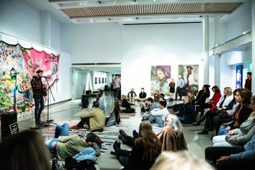 A diverse audience sits on the floor of an art gallery, listening to a speaker in front of colorful artwork.