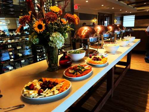 A buffet table with various dishes, including fruits, salads, and pastries, surrounded by flowers and wine bottles.