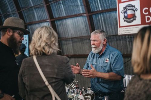 A bearded man in a blue shirt gestures while speaking to two people at a bar event in a rustic setting.