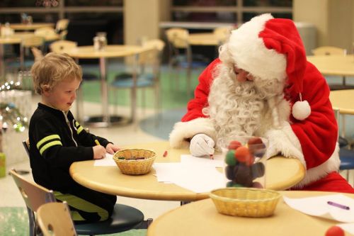 A child and Santa Claus sit at a table, drawing with crayons and paper in a festive setting.