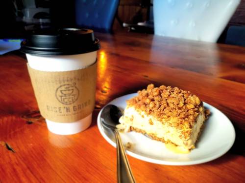 A slice of dessert with a crumbly topping on a plate next to a coffee cup on a wooden table.