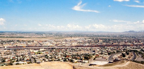 A panoramic view of a cityscape with buildings, roads, and mountains under a blue sky with scattered clouds.